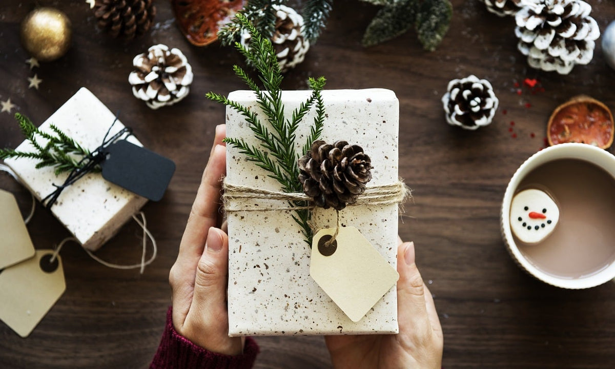 Hands holding a small wrapped Christmas gift with a pine cone next to a mug of cocoa